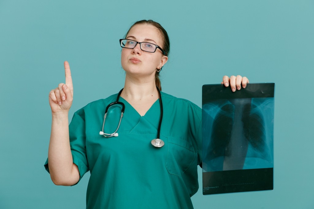 young woman nurse medical uniform with stethoscope around neck holding lung xray looking confident showing index finger having good news standing blue background